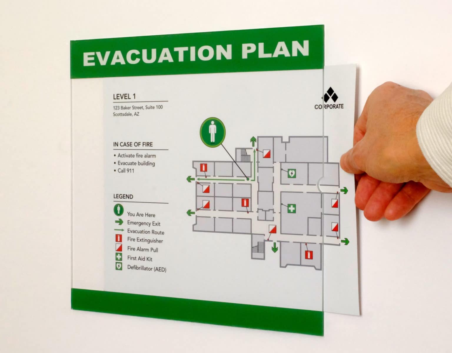 Hand holding printed evacuation plan with green header showing level 1 office layout, fire safety icons, AED location, and emergency procedures