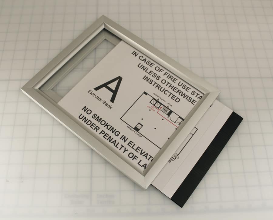 Printed evacuation sign sliding out of the top of an aluminum slide-in frame, showing how inserts are changed without removing the frame from the wall